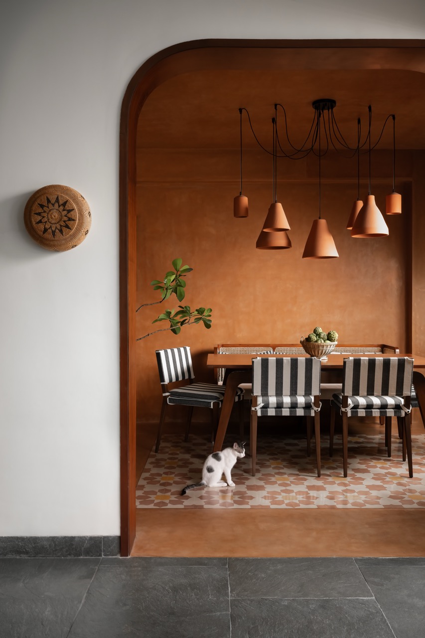 The dining room has terracotta-coloured walls and ceiling with accent flooring by Bharat Floorings & Tiles. A deep, rounded archway frames the space, while the table by Urban Ladder, chairs by Josmo, bench by ma+rs, and terracotta light cluster by Ara Living finish the look