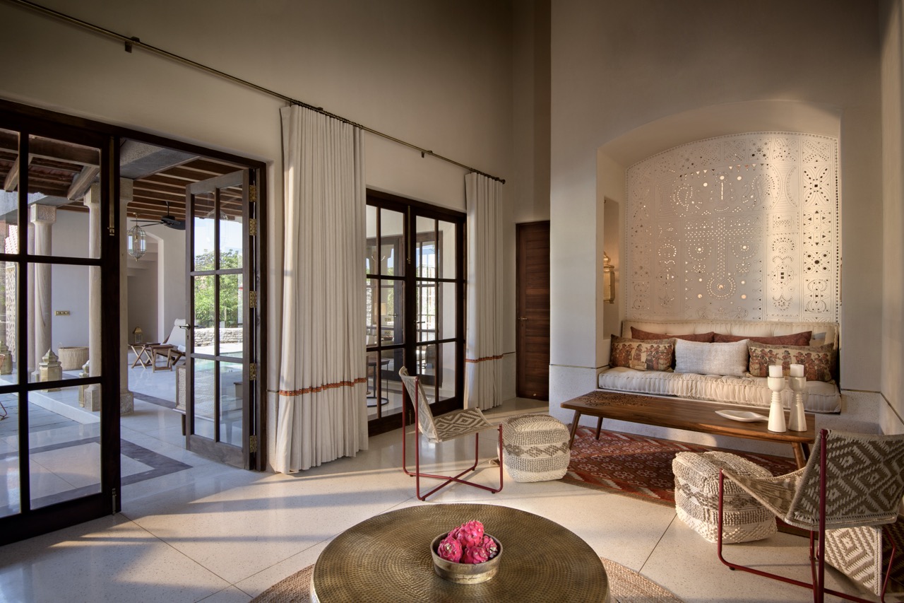 In this villa, a built-in seating niche sits against a panel of white Gujarati lippan work, while terrazzo floors and open doors extend the room into the courtyard beyond