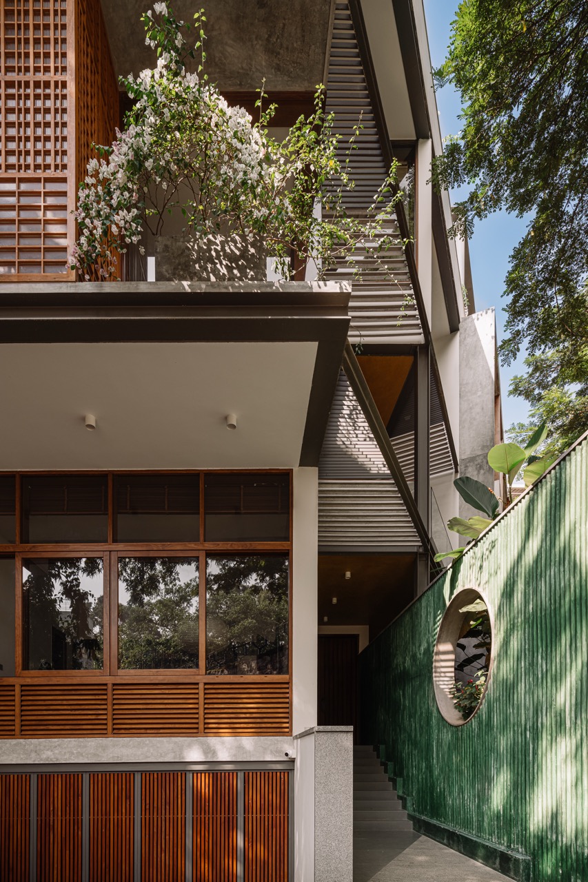 The entrance is framed by a green corrugated concrete wall and exposed precast pergolas, guiding movement toward the main door. Woodwork by RitikaaWood adds warmth to the concrete setting