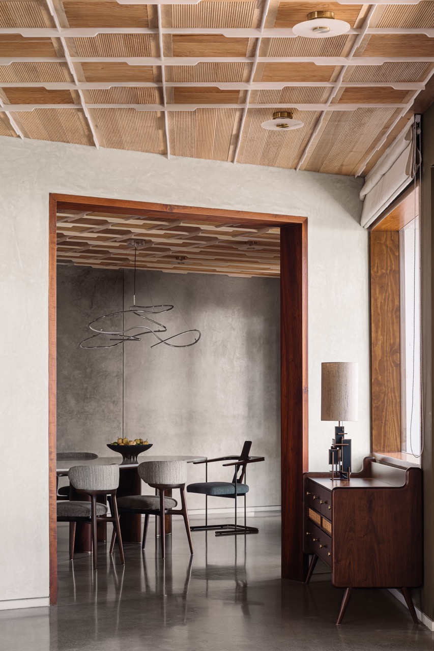 In the dining area, coffered fluted wooden ceiling offsets the grey oxide walls and flooring, with a lamp on the side console by Phantom Hands adding a soft accent
