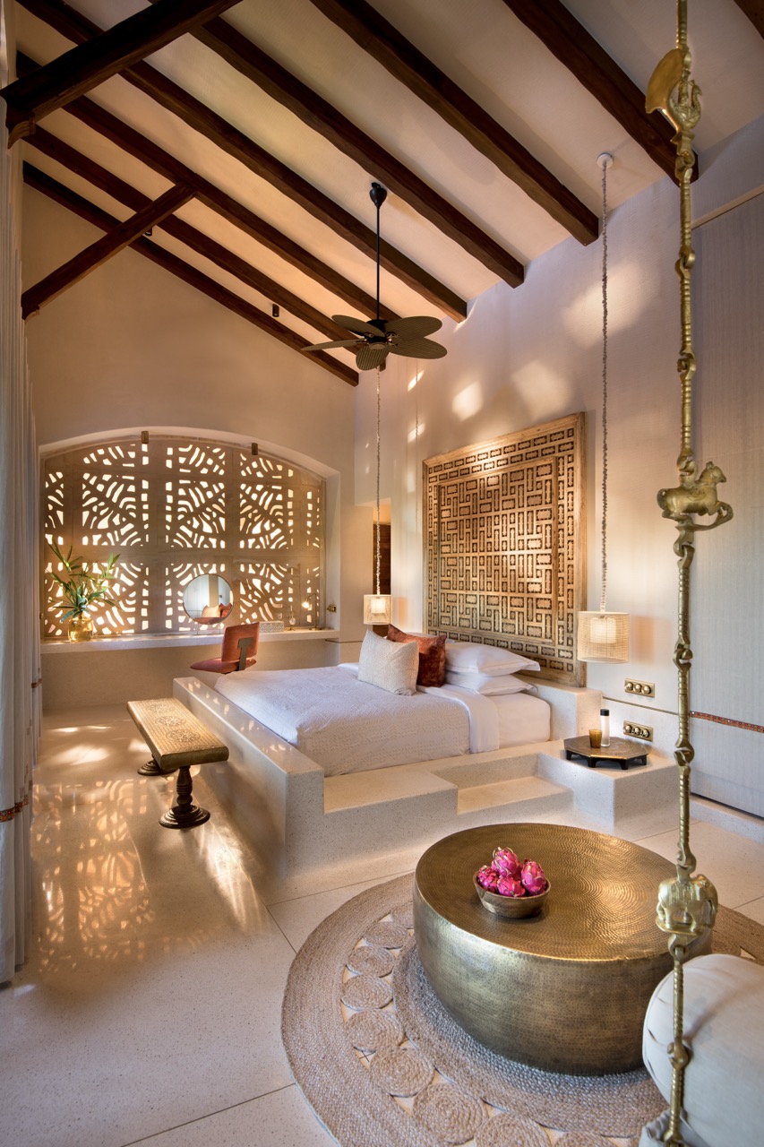 A raised terrazzo bed anchors this luxurious room, set against a carved panel, with filtered light from the jali casting shifting patterns across the floor and walls